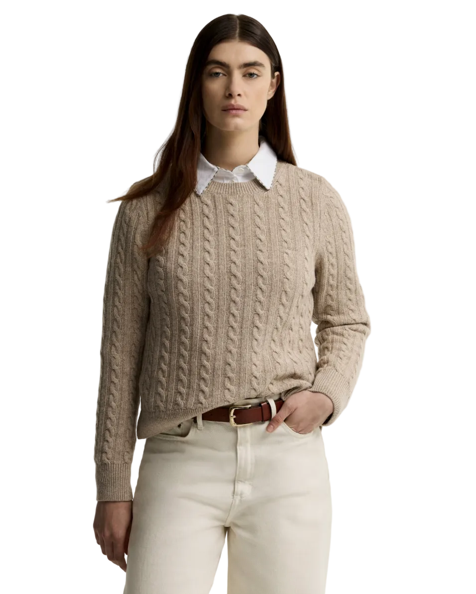 A woman modeling a beige cable-knit crewneck sweater over a white collared shirt, paired with cream trousers and a brown belt, against a plain white studio background.