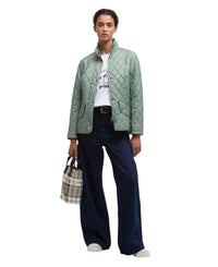 A model stands against a light gray studio background wearing a sage green quilted jacket over a white graphic T-shirt, dark navy wide-leg trousers, and white shoes, while holding a beige plaid tote bag.