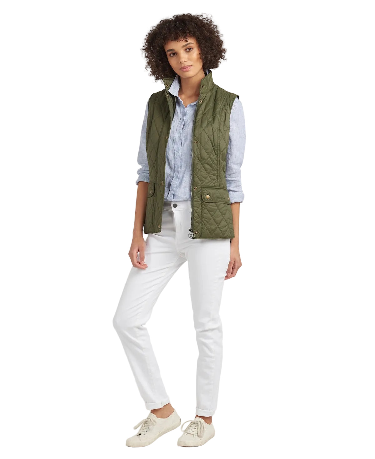 A woman modeling an olive green quilted sleeveless vest over a light blue button-up shirt, paired with white pants and cream sneakers, against a plain white studio background.
