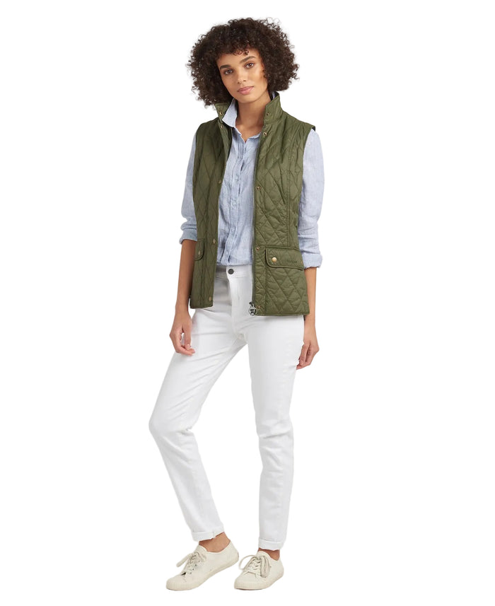 A woman modeling an olive green quilted sleeveless vest over a light blue button-up shirt, paired with white pants and cream sneakers, against a plain white studio background.