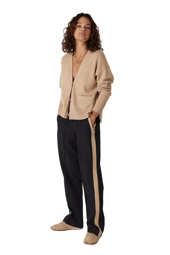 A woman stands against a plain light background wearing a beige cardigan, dark navy wide-leg trousers with beige side stripes, and beige slip-on shoes.