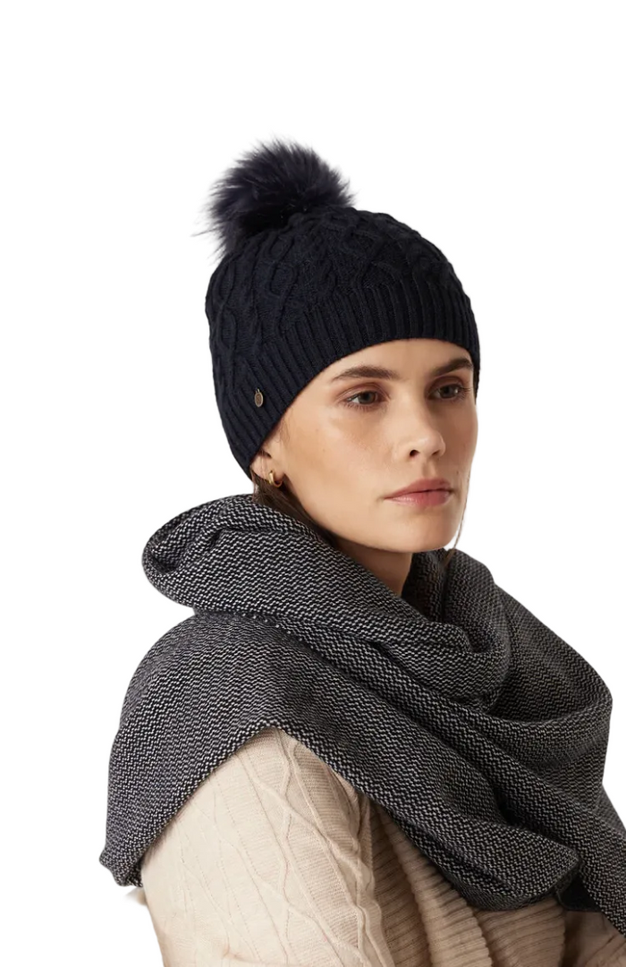 Person wearing a black knit beanie with a pom-pom and a dark gray scarf on a light background