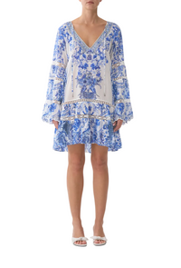 A woman modeling a blue and white toile-patterned long-sleeve mini dress with a V-neckline, ruffled hem, and sheer lace detailing on the sleeves, paired with white heeled sandals against a light gray studio background.