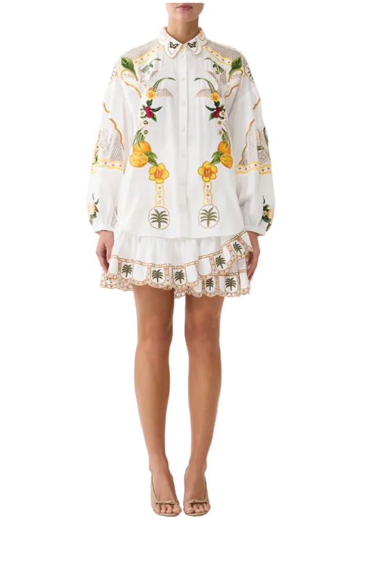 A model wearing a white embroidered long-sleeve blouse and matching mini skirt set. The white fabric is heavily decorated with yellow, green, and red floral and botanical embroidery, finished with a ruffled lace hem.