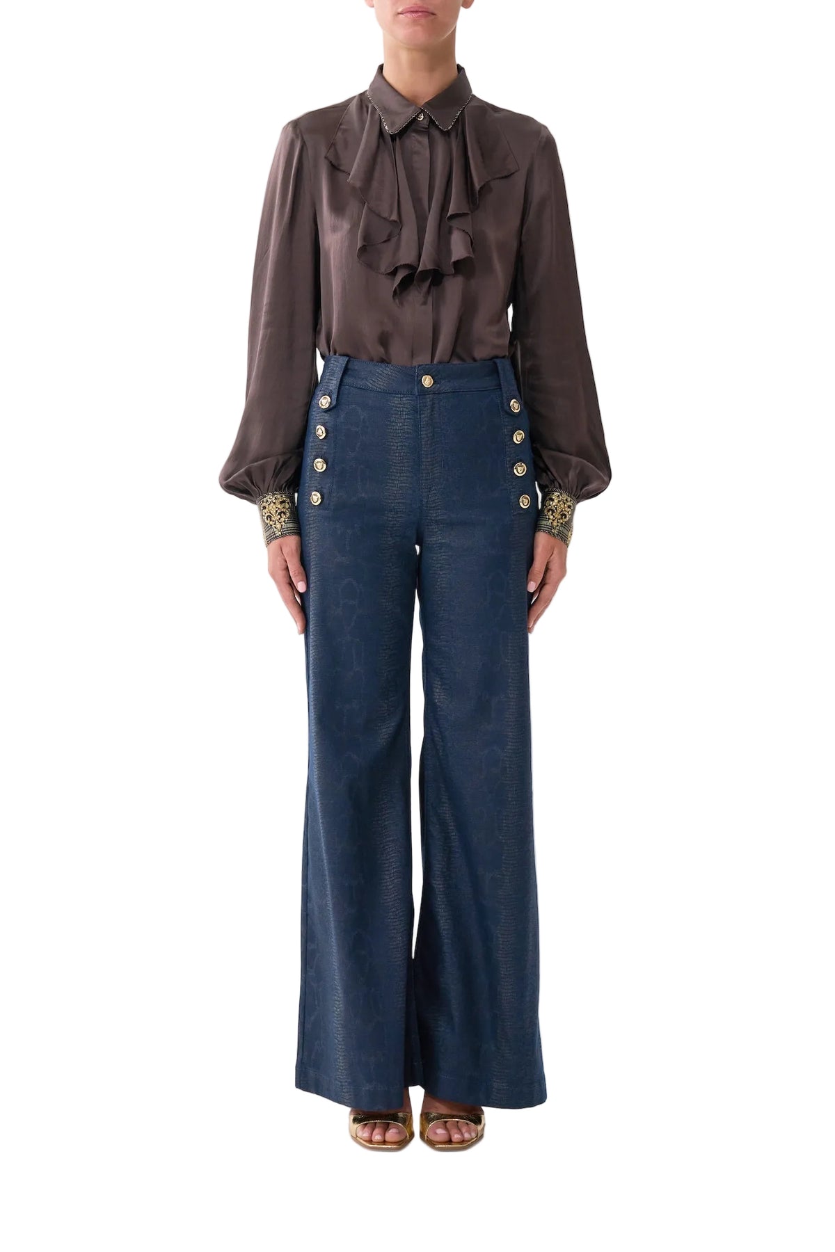 A model wearing a dark brown long-sleeve blouse with a ruffled tie-neck and gold-embellished cuffs, paired with high-waisted blue wide-leg trousers featuring gold buttons down the sides, set against a plain light gray background.