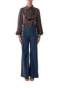 A model wearing a dark brown long-sleeve blouse with a ruffled tie-neck and gold-embellished cuffs, paired with high-waisted blue wide-leg trousers featuring gold buttons down the sides, set against a plain light gray background.