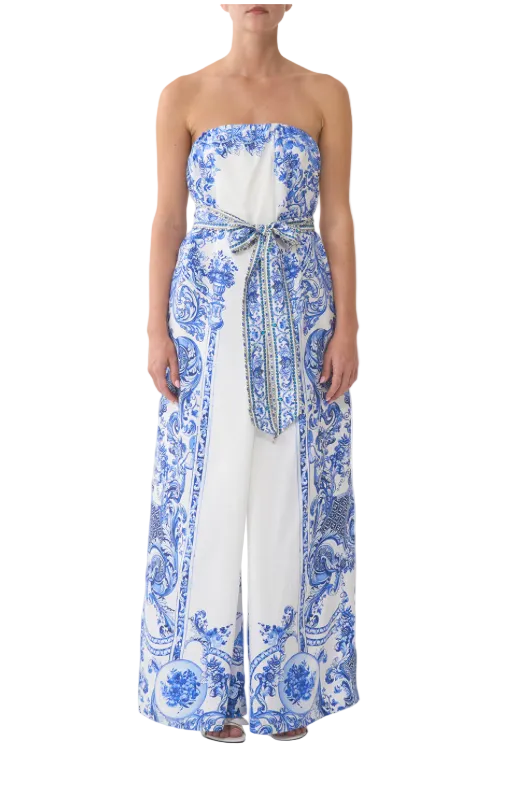 A strapless white wide-leg jumpsuit with a blue and white baroque pattern, featuring a tie-waist sash, modeled against a light gray background.