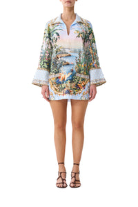 A model wearing a long-sleeve mini shirt dress with a colorful tropical landscape print, featuring a light blue base with green foliage, a beach scene, and gold ornamental borders. The dress has a split neckline and flared cuffs, paired with brown strappy high-heeled sandals against a plain white background.