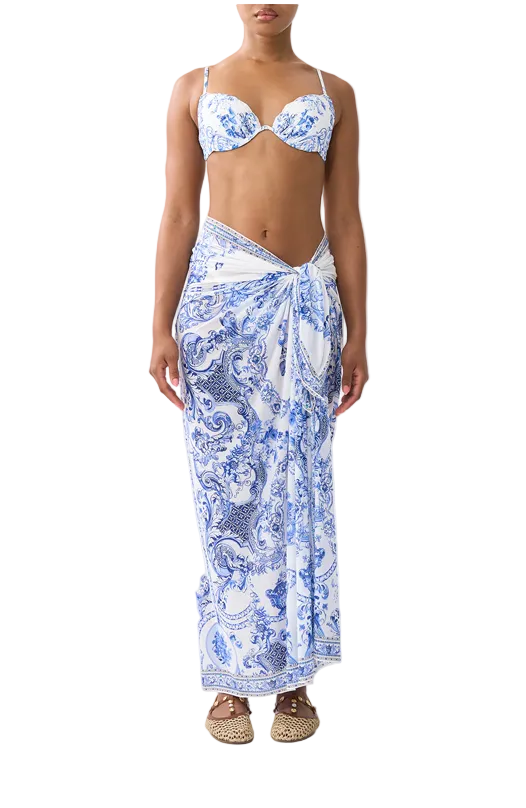 A model wearing a two-piece swim set and a matching sarong with a blue and white paisley pattern, paired with brown woven sandals, against a plain light gray background.