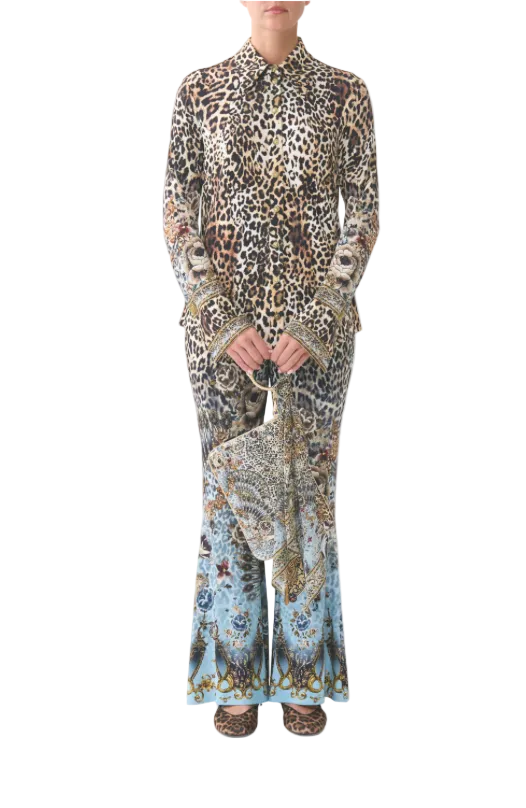 A model wearing a matching long-sleeve button-up shirt and wide-leg trousers set in a brown, beige, and black leopard print, with a matching leopard-print handbag and leopard-print shoes, against a light gray background.