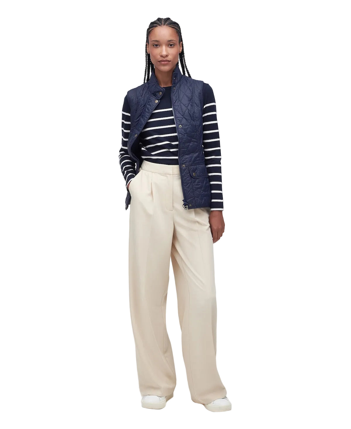 A model stands against a plain light gray studio background wearing a navy quilted vest over a navy and white striped long-sleeve top, paired with cream wide-leg trousers and white shoes.