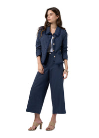 A woman modeling a navy blue two-piece suit consisting of a cropped jacket with ruffled trim and matching wide-leg cropped pants, styled with beige heeled sandals against a light gray studio background.