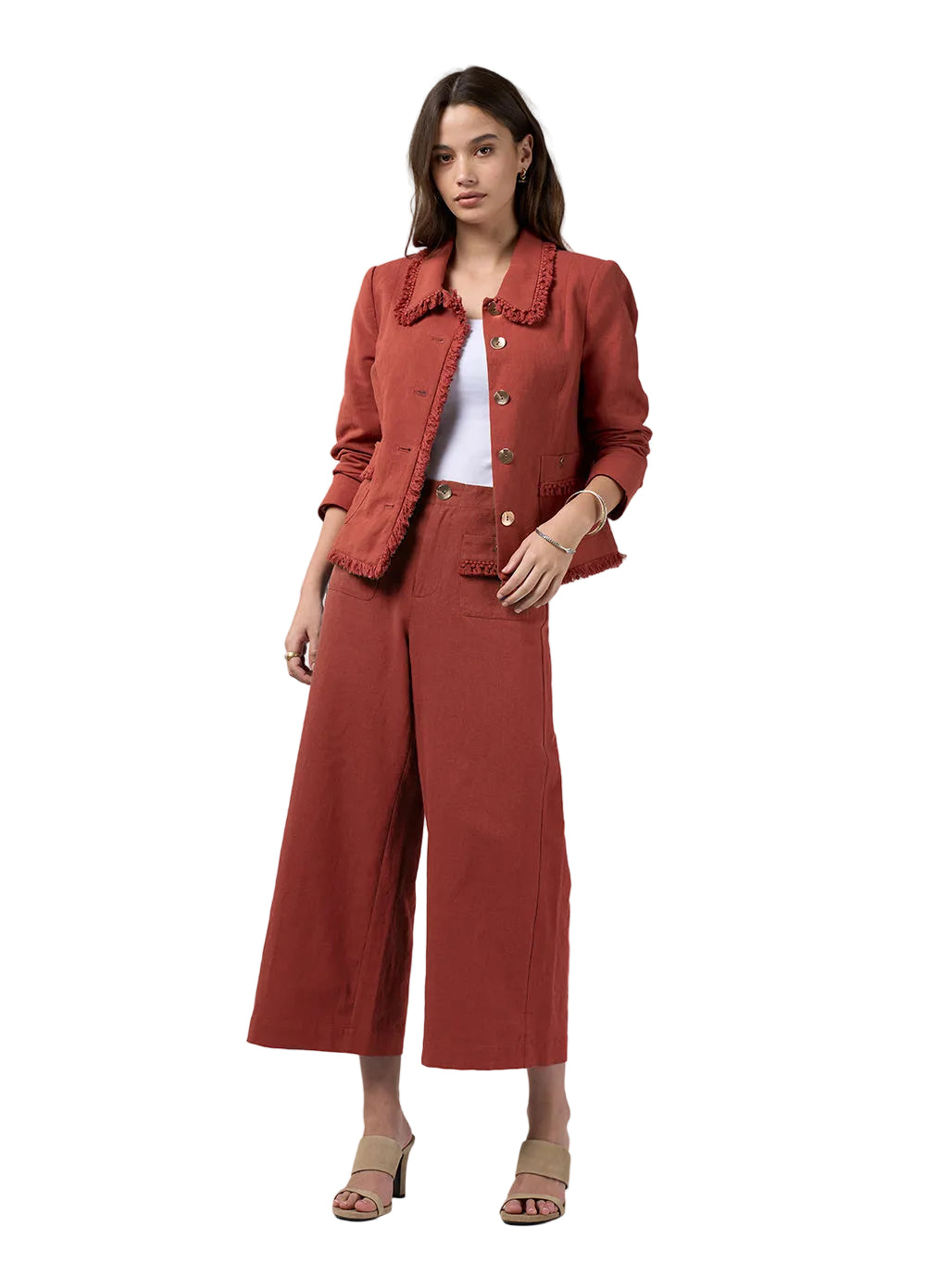 A woman modeling a rust-colored two-piece set consisting of a cropped jacket with ruffle trim and matching wide-leg cropped pants, styled with a white top and beige heeled sandals against a light gray studio background.