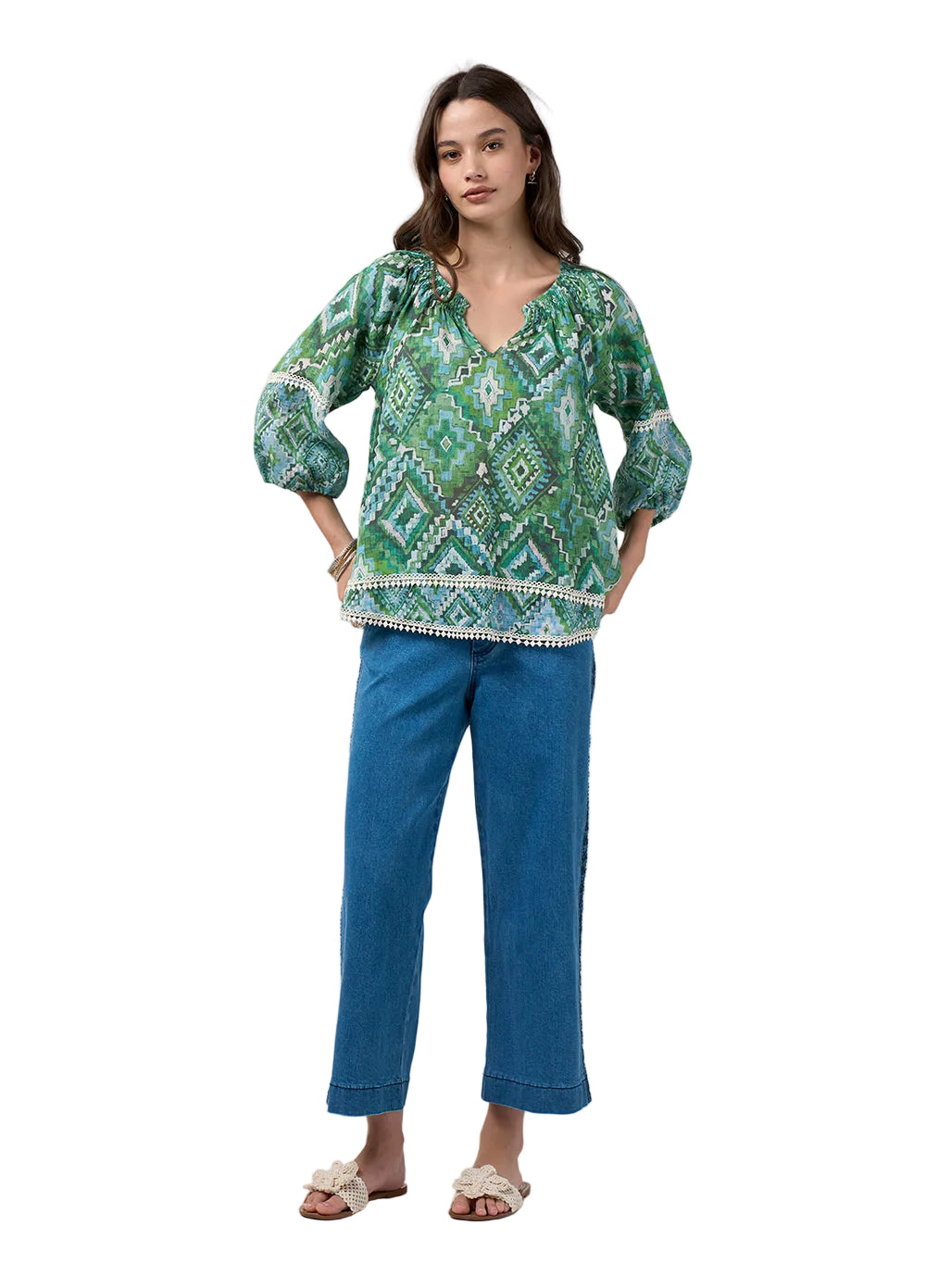 A woman modeling a green and white patterned bohemian-style blouse with puff sleeves and a tassel hem, paired with blue jeans and beige slide sandals, against a light gray studio background.