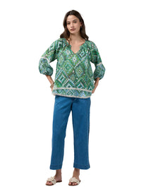 A woman modeling a green and white patterned bohemian-style blouse with puff sleeves and a tassel hem, paired with blue jeans and beige slide sandals, against a light gray studio background.