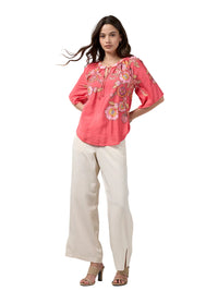 A woman modeling a coral pink short-sleeve blouse with colorful floral embroidery on the chest, paired with cream wide-leg trousers and beige heeled sandals, against a light gray studio background.