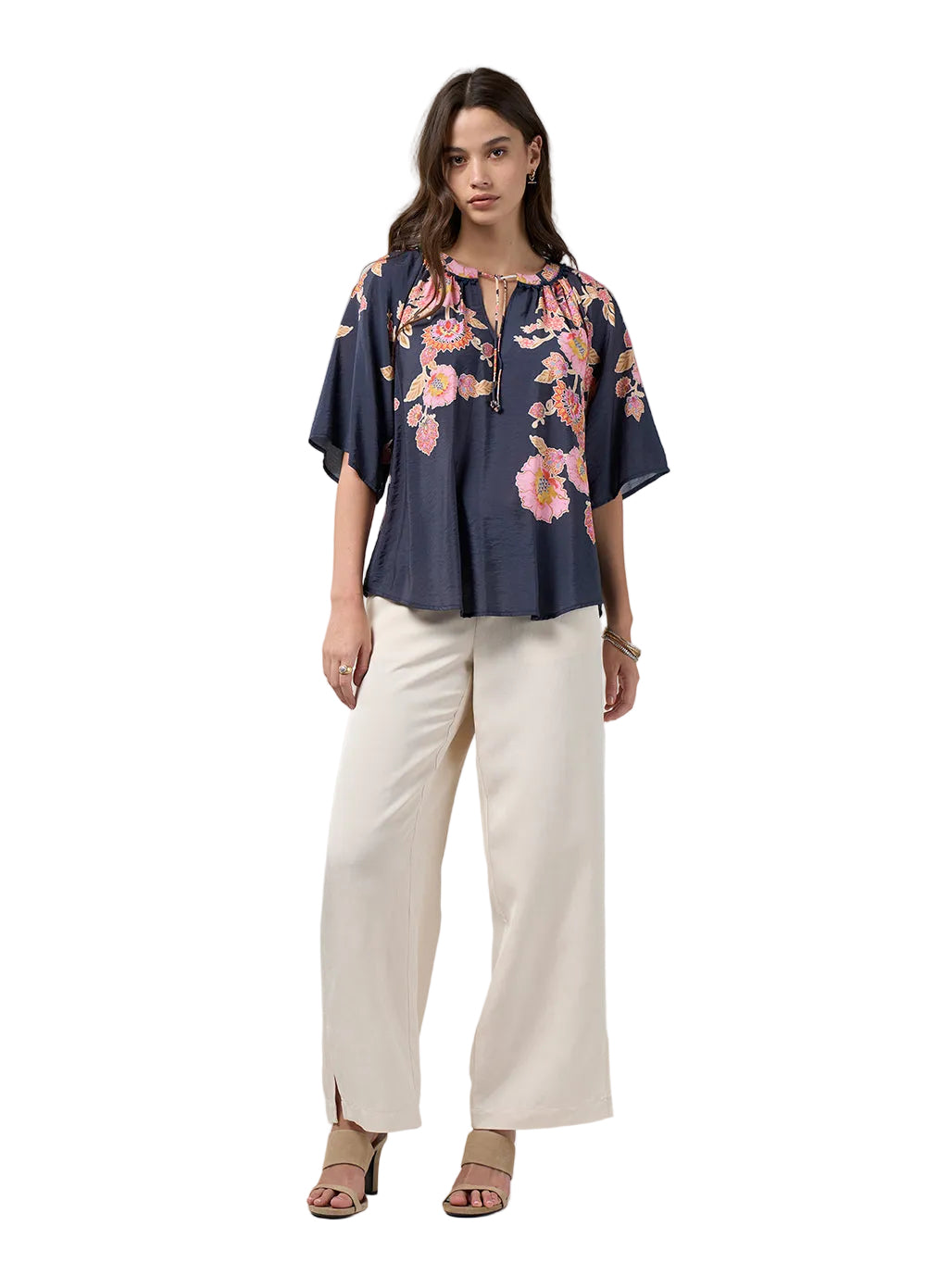 A woman modeling a navy blue short-sleeve blouse with a pink floral print and cream wide-leg pants against a light gray studio background.