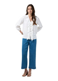 A woman modeling a white long-sleeve blouse with ruffle details down the front and blue wide-leg cropped jeans, standing against a light gray studio background.
