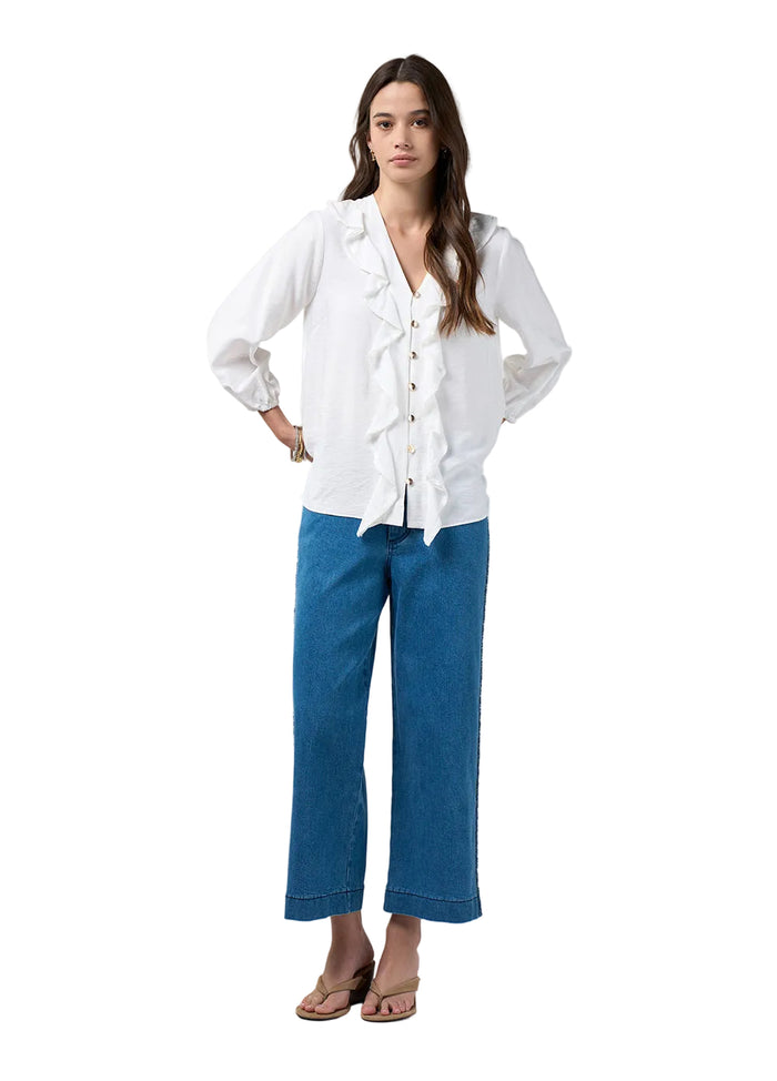 A woman modeling a white long-sleeve blouse with ruffle details down the front and blue wide-leg cropped jeans, standing against a light gray studio background.