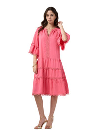 A woman modeling a tiered pink dress with ruffled sleeves and lace trim, paired with gold heeled sandals, against a light gray studio background.