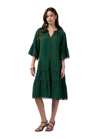 A woman modeling a dark green tiered midi dress with three-quarter bell sleeves and lace trim, paired with black strappy sandals against a light gray studio background.