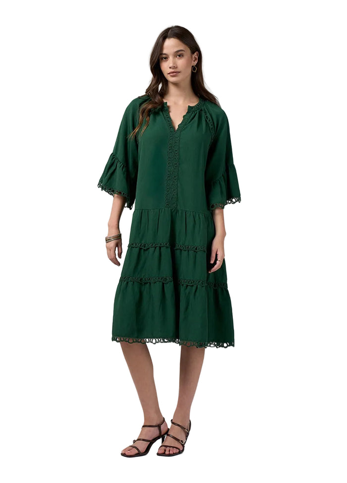A woman modeling a dark green tiered midi dress with three-quarter bell sleeves and lace trim, paired with black strappy sandals against a light gray studio background.