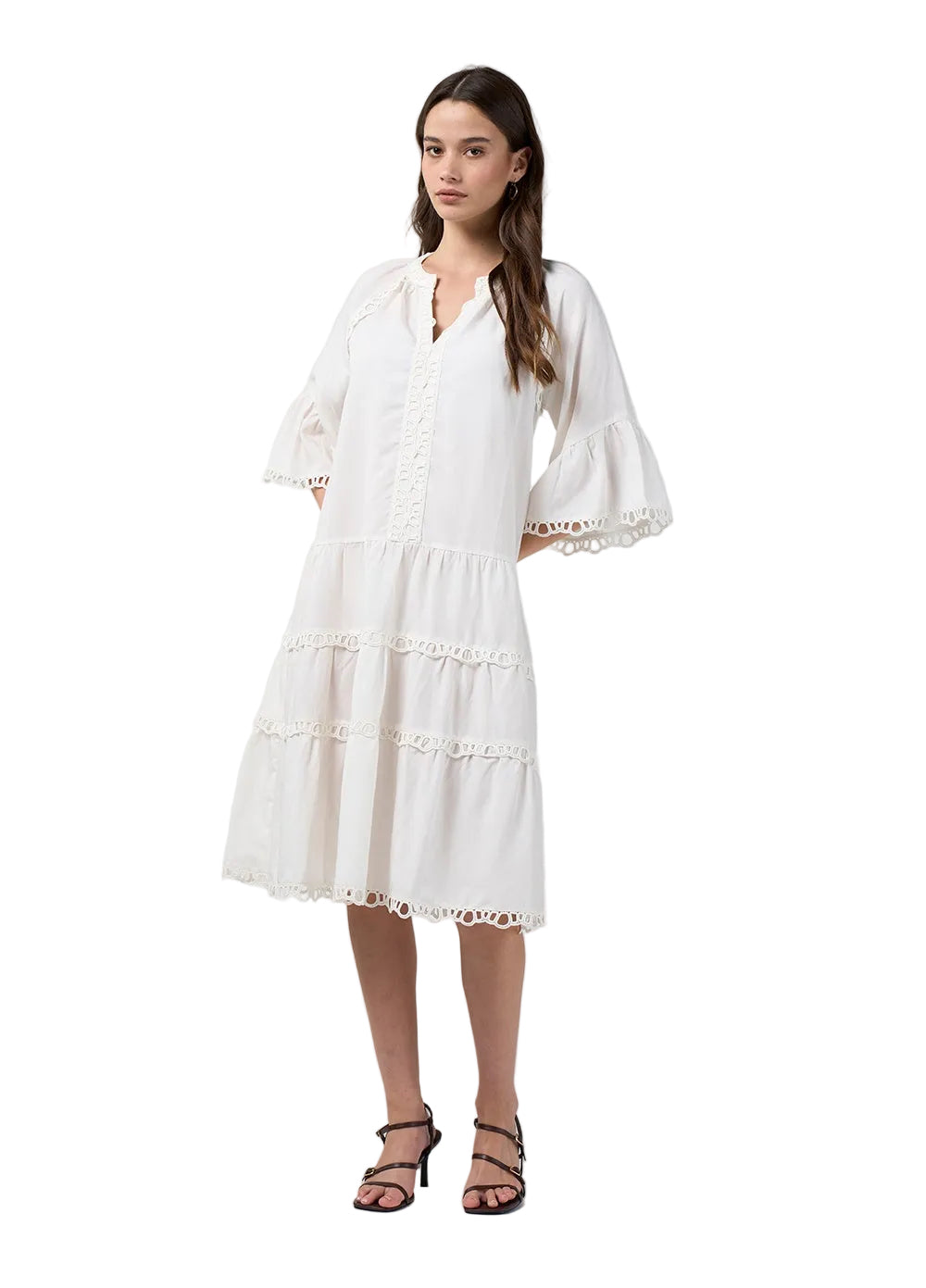 A woman modeling a white tiered midi dress with wide flutter sleeves and eyelet lace trim, paired with black strappy heeled sandals, against a light gray studio background.