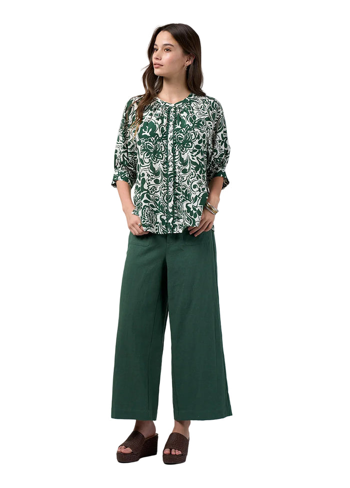 A woman modeling a green and white patterned blouse with three-quarter sleeves, paired with dark green wide-leg pants and brown wedge sandals, against a light gray studio background.