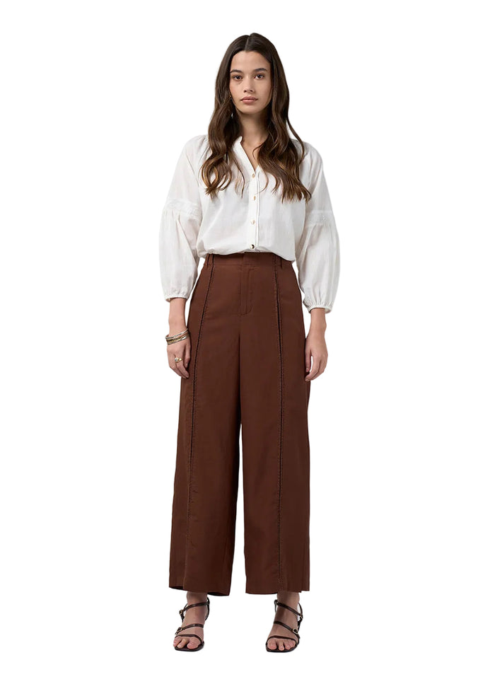 A woman modeling a white long-sleeve blouse tucked into high-waisted, wide-leg brown trousers, paired with black strappy high-heeled sandals, against a light gray studio background.