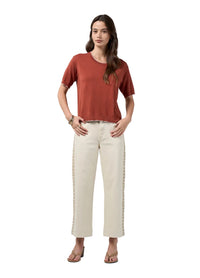 A woman modeling a rust-colored short-sleeve t-shirt and off-white cropped pants with decorative side detailing, standing against a light gray studio background.
