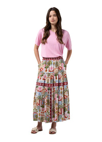 A woman modeling a pink short-sleeve t-shirt and a long, tiered maxi skirt with a colorful tropical and beach-themed print, standing against a light gray studio background.