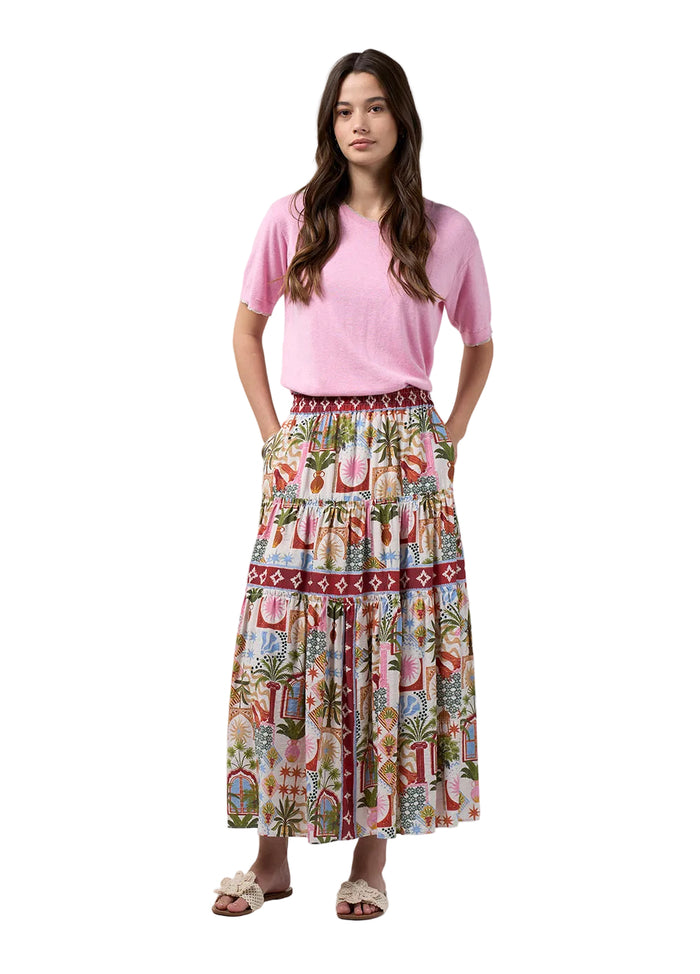 A woman modeling a pink short-sleeve t-shirt and a long, tiered maxi skirt with a colorful tropical and beach-themed print, standing against a light gray studio background.
