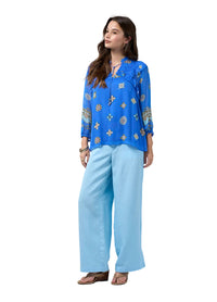 A woman modeling a bright blue long-sleeve blouse with a bohemian-style multicolor print and light blue wide-leg pants, standing against a neutral light gray studio background.