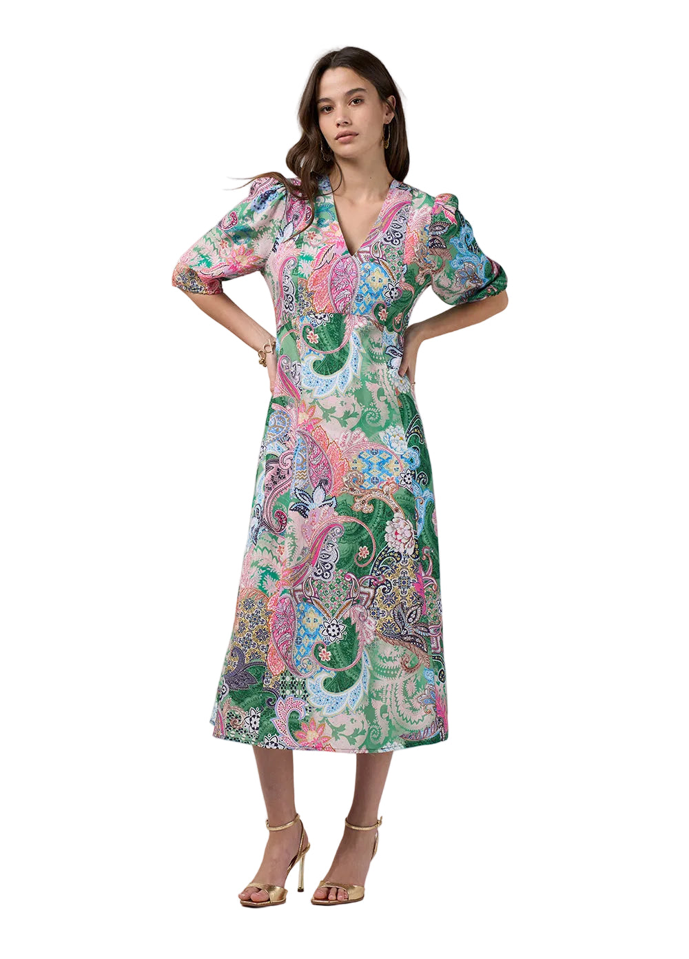 A woman modeling a midi wrap dress with puff sleeves and a vibrant paisley print in green, pink, blue, and white, paired with gold high-heeled sandals against a light gray studio background.