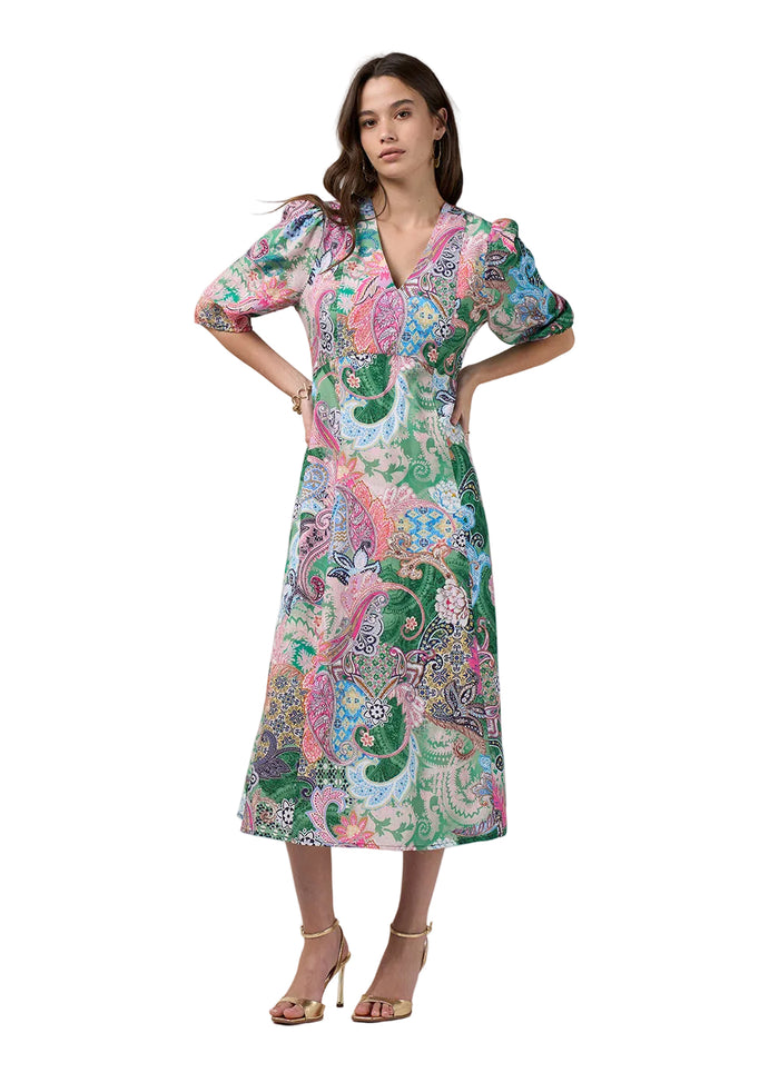A woman modeling a midi wrap dress with puff sleeves and a vibrant paisley print in green, pink, blue, and white, paired with gold high-heeled sandals against a light gray studio background.
