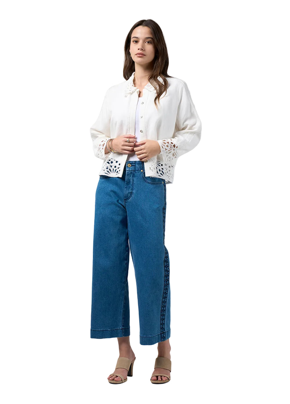 A woman modeling a white long-sleeve blouse with delicate lace cutouts on the sleeves and hem, paired with blue cropped jeans and beige heeled sandals, against a light gray studio background.