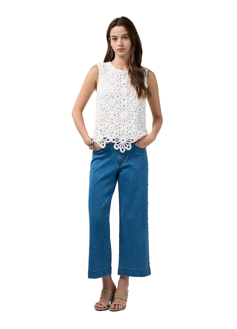 A woman modeling a sleeveless white crochet top and blue cropped jeans against a light gray studio background.