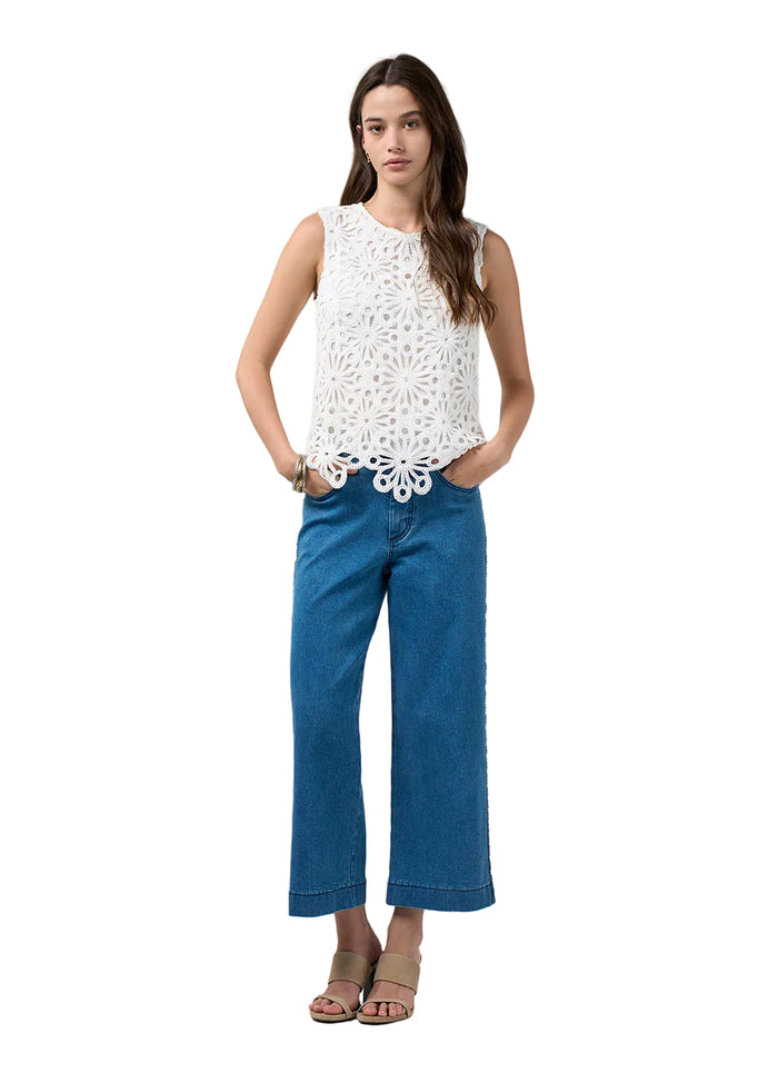 A woman modeling a sleeveless white crochet top and blue cropped jeans against a light gray studio background.