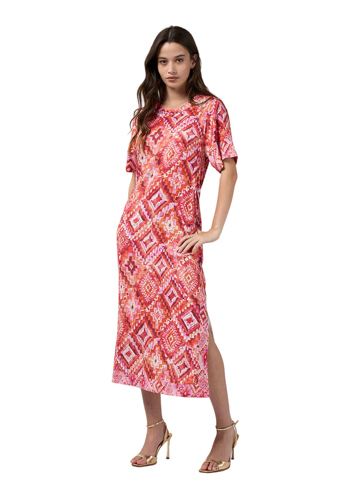 A woman modeling a short-sleeve midi dress with a vibrant red, pink, and orange geometric pattern, paired with gold high-heeled sandals, against a light gray studio background.