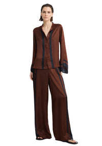 A model wearing a matching two-piece lounge set in dark brown satin with navy blue stripe accents, consisting of a long-sleeve button-up shirt and wide-leg pants, photographed against a light gray studio background.