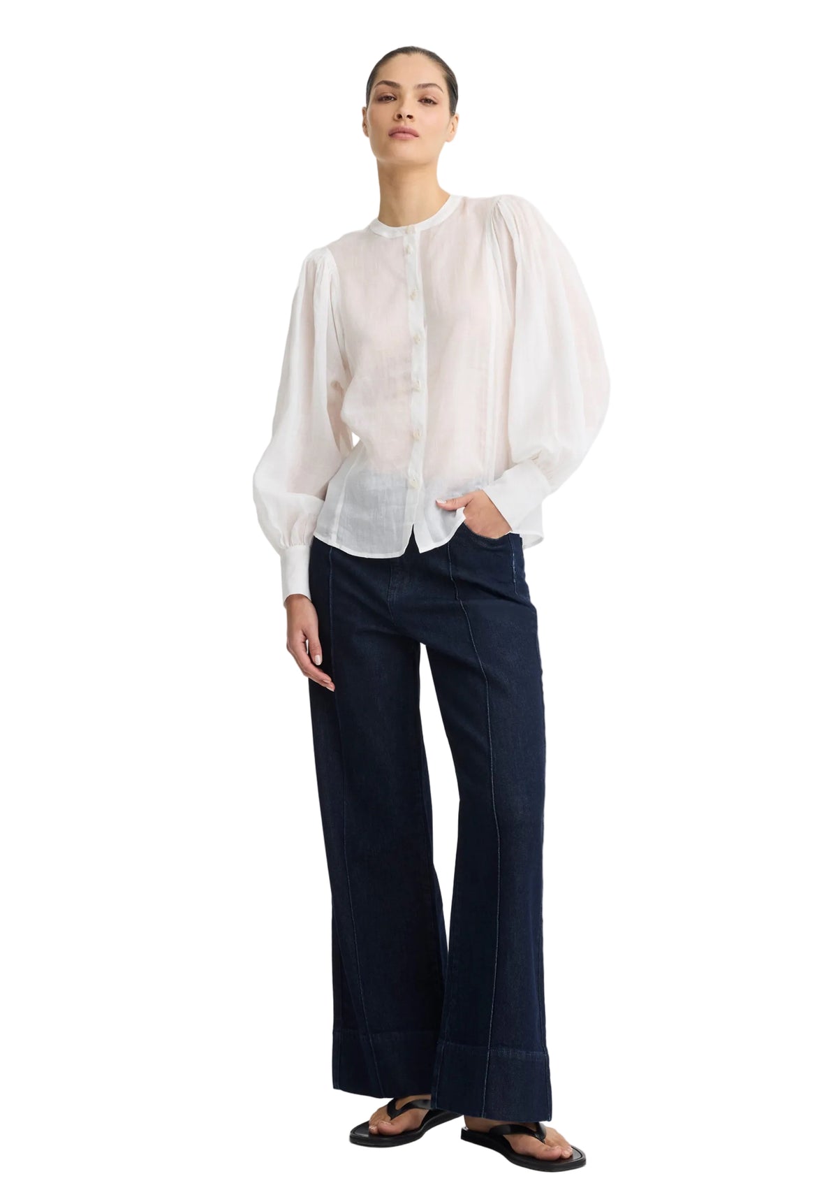 A model stands against a plain white studio background wearing a sheer white long-sleeve blouse with dramatic puffed shoulders and dark navy wide-leg trousers.