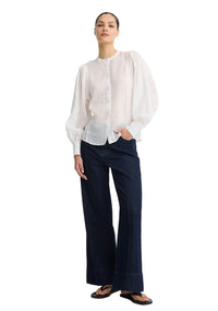 A model stands against a plain white studio background wearing a sheer white long-sleeve blouse with dramatic puffed shoulders and dark navy wide-leg trousers.