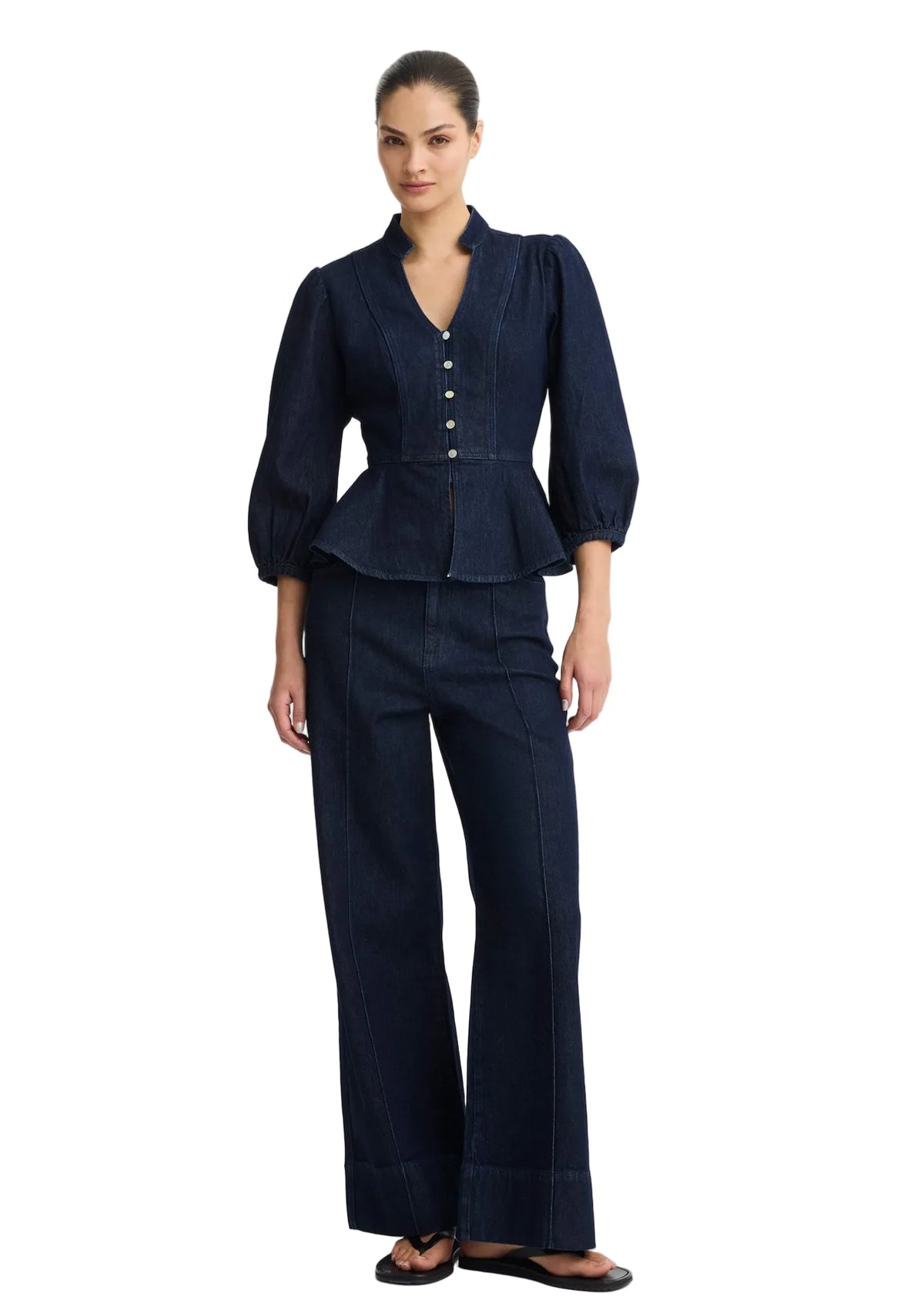 A model wearing a dark indigo denim two-piece set consisting of a long-sleeve peplum blouse with button details and matching wide-leg trousers, photographed against a solid light gray background.