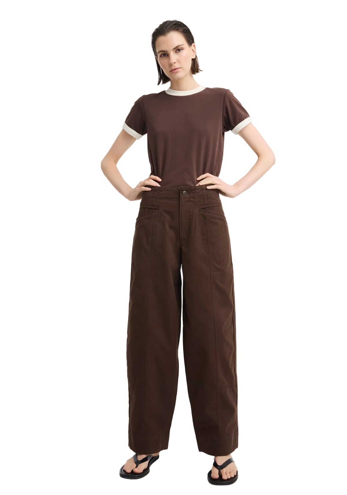 A model stands against a light gray studio background wearing a dark brown short-sleeve t-shirt with white ribbed trim at the collar and sleeve cuffs, paired with matching dark brown wide-leg trousers and black sandals.