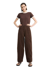 A model stands against a light gray studio background wearing a dark brown short-sleeve t-shirt with white ribbed trim at the collar and sleeve cuffs, paired with matching dark brown wide-leg trousers and black sandals.