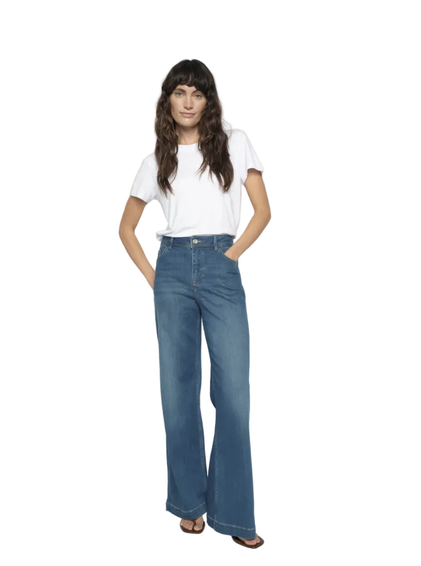 A woman modeling a pair of high-waisted, wide-leg blue jeans with a white t-shirt, standing against a light gray studio background.