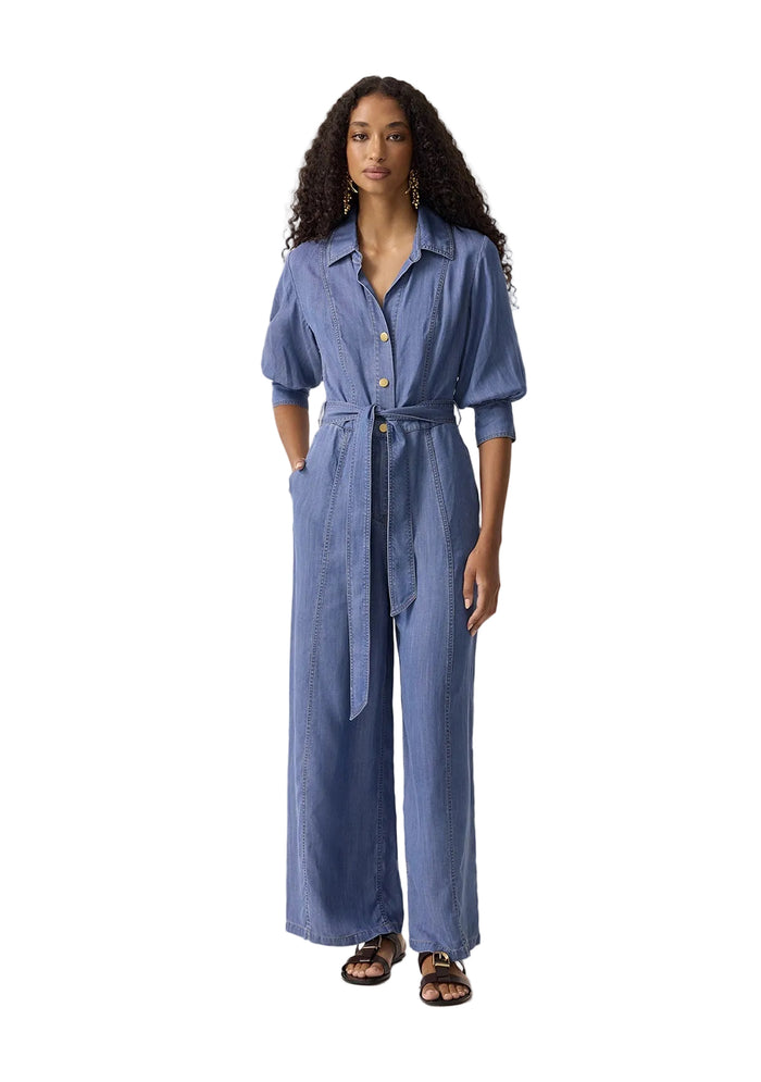 A woman modeling a blue denim-like shirtdress with short sleeves, a button front, a self-tie belt, and wide legs, standing against a plain light beige studio background.
