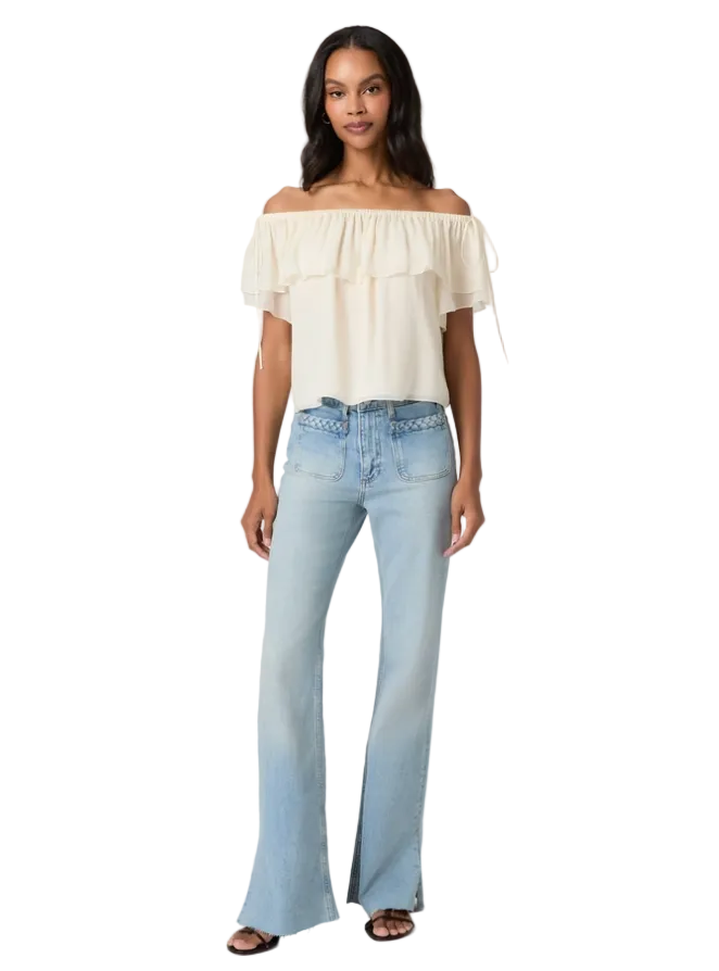 A model stands against a light gray studio background wearing an off-the-shoulder cream ruffled blouse and light-wash blue flare jeans with decorative silver studs on the hips.