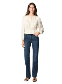 A woman modeling a cream-colored long-sleeve blouse with a split neckline and gathered waist, paired with dark blue jeans and brown sandals, against a plain white background.