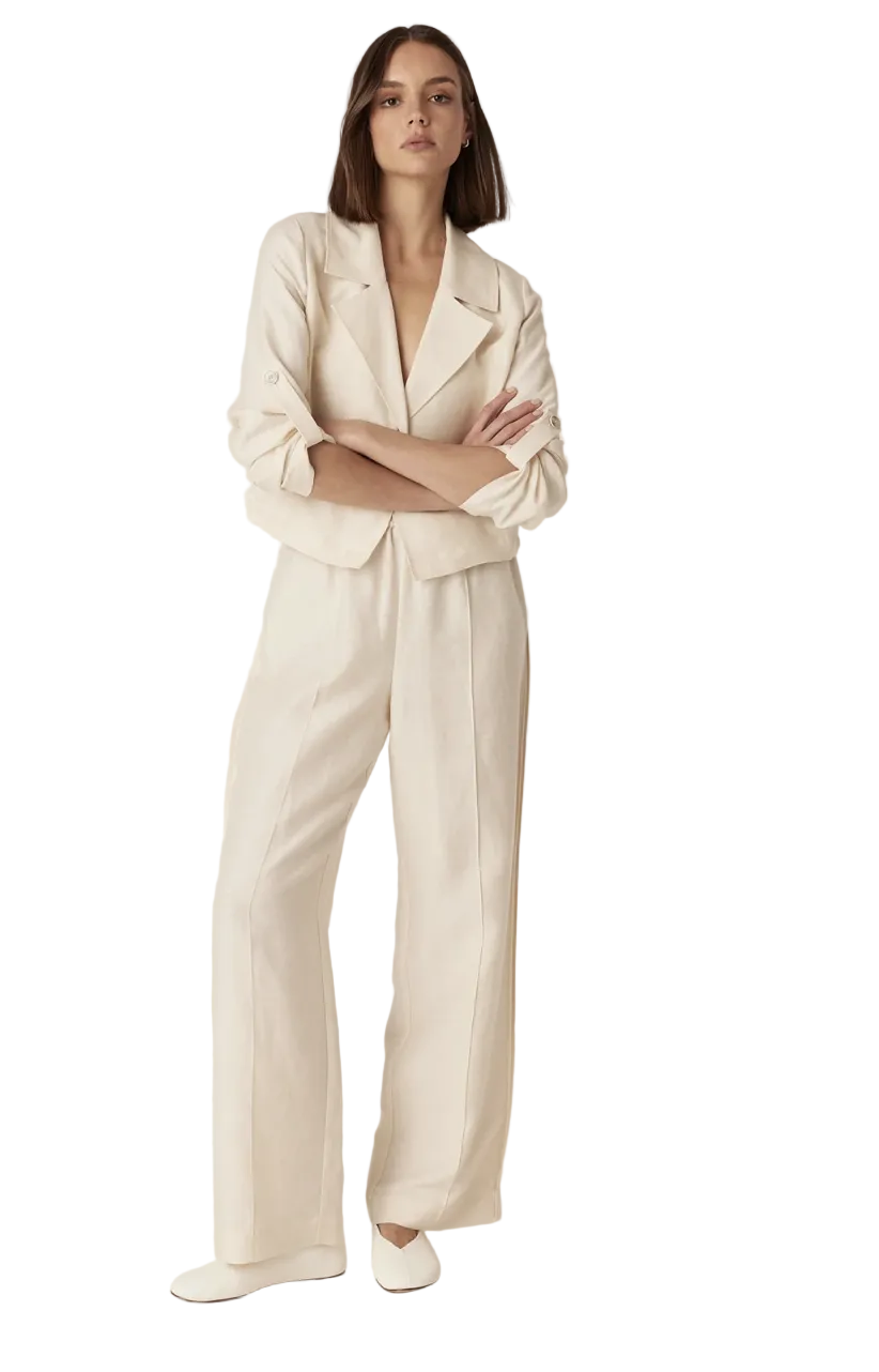 A woman stands against a plain light beige studio background wearing a matching cream-colored tailored set consisting of a cropped blazer and wide-leg trousers, paired with white slip-on shoes.